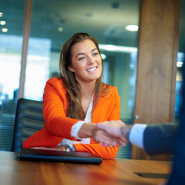 A white woman leans across a desk to shake a man’s hand