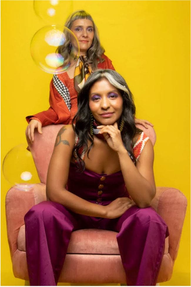 A bright yellow background with two women age 40+ wearing funky clothes looking directly into he camera