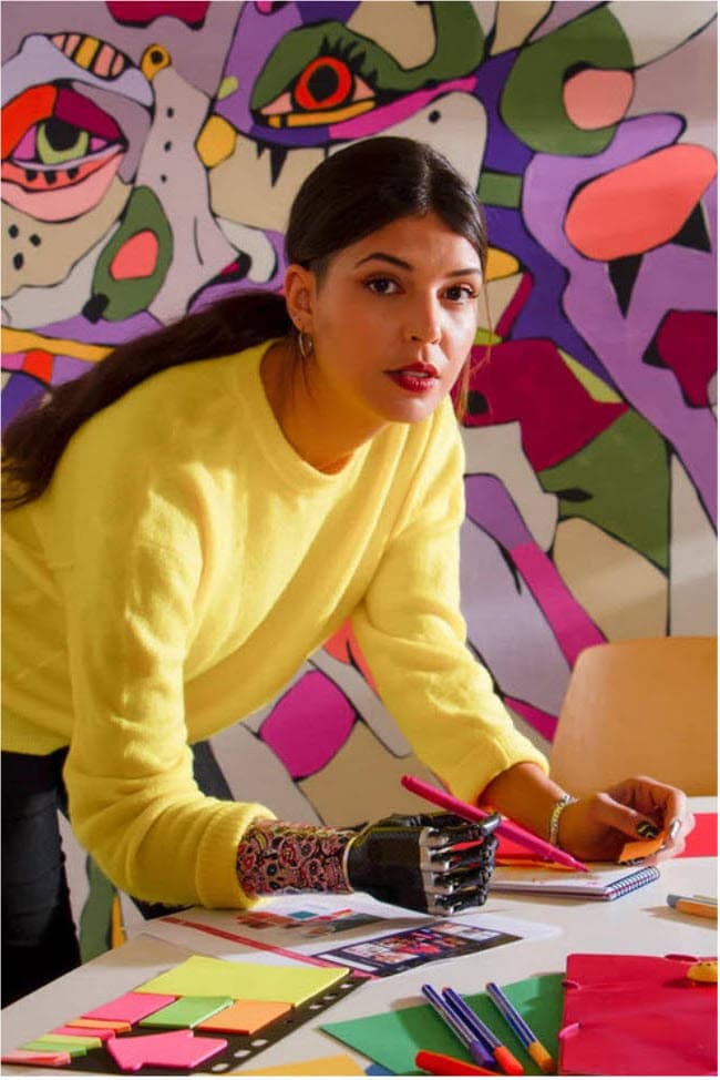 A young white woman in her 20s is in an art studio with a multi-coloured background drawing using her prosthetic arm